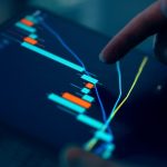 A finger pointing at a digital trading chart on a tablet screen with colorful candlestick graphs